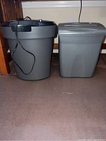 Two gray paper shredders on floor under desk, one taller cylindrical bin style, one shorter rectangular bin style.