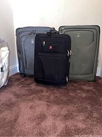 Three suitcases standing on carpet against a wall: black Swissgear in front, two Travelpro suitcases behind in gray and green.