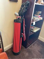Red vintage style golf bag standing next to bookshelf in office setting, showing full bag with golf clubs inside.