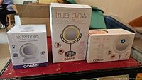 Three boxed lighted vanity mirrors by Conair on a red table: two True Glow models and one Reflections model.
