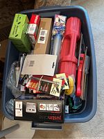 View of assortment of tools and gadgets mostly in packaging inside a plastic bin, including a red handled tool case and various small packages.