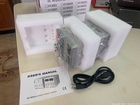 Image of the DC regulated power supply unit in foam packaging with power cord and user's manual on a table.