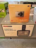 Photo showing the Canon PowerShot A2300 camera box placed on top of the Crosley Audiophile AM/FM Receiver box on a table, demonstrating the lot contents.