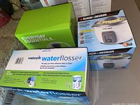 Photo showing boxed items: Everyday Essentials green box, Black & Decker rice cooker box, and Waterpik water flosser box stacked together on floor.