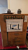 Box labeled U-Haul with a printed picture and toner text stating Samsung 1000 Watt 3D Blu-ray Home Theatre System.