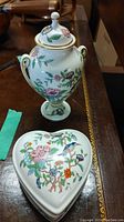 Photo showing urn with lid and heart-shaped trinket box with floral and bird decorations on wooden surface.