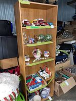 Full view of pressboard bookshelf containing various items on shelves including toy vehicles, decorative glass balls, ceramic flower arrangements, and crafting supplies.