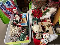 Two large plastic bins filled with assorted Christmas decorations, including string lights, bead garlands, Santa figures, table cloth, and a plush teddy bear.