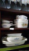 Shelf with stacked Corning Ware Blue Cornflower dishes including casserole and lasagna dishes, plus a square casserole with vegetable design.