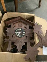 Clock components in cardboard box: wooden cuckoo clock body with carved leaf and bird details, clock face with Roman numerals, additional wooden carved pieces, and metal weights.