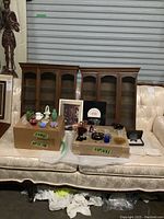 Two wooden curio cabinets with glass doors and shelves, displayed on a cream color upholstered sofa. Various glass miniatures are placed in front and on cardboard boxes labeled 'fragile'.