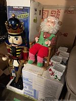 View of wooden nutcracker standing next to rocking Santa doll on boxes, including partial view of white decorative plant holders
