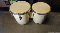 Pair of connected bongo drums with cream-colored shells and white drumheads on a dark surface, showing overall condition and construction.