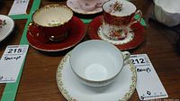 Three teacups and saucers including a red set with gold trim and floral detailing, and a white set with floral border