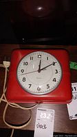 Front view of the vintage WestClox electric clock showing red square casing, clock face with Arabic numerals, metal hands and attached cord and plug.