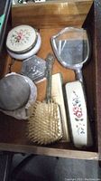 View of the full set inside a drawer showing two brushes with floral covers, comb, powder jar with floral lid, metal ring box and hand mirror.