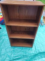 Photo of a medium brown wood grain laminate bookcase with three shelves, photographed outdoors on a blue tarp.