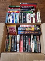 Top view of two boxes filled with used adult suspense, thriller, and crime books arranged upright with titles visible on spines.