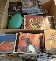 Top view of box filled with stacked CDs and a green football shaped item to show the quantity and condition of the lot.