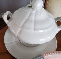 White ceramic soup tureen with matching warmer plate, featuring double handles and a lid with a knob.