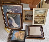Multiple miscellaneous framed prints and paintings arranged leaning on a carpeted floor, featuring waterfall, autumn trees, bridges and other natural scenes.