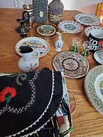 Overview of lot showing assortment of vintage decorative plates, pitchers, mugs, candlesticks, and textiles arranged on a wooden table.