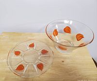 Large clear glass punch/fruit bowl with orange leaf decoration, and matching round glass plate with orange leaf design on wooden surface.
