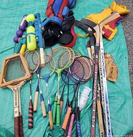 All items laid out on a blue tarp including badminton and tennis rackets, birdies, hockey sticks, baseball glove, two super soakers, and youth life jacket. Shows overall lot contents and condition.