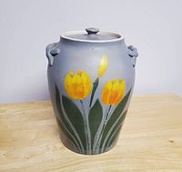 Front view of large stoneware cookie jar with hand-painted yellow tulips on grayish beige surface, placed on wood surface.
