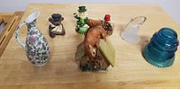 Vintage figurines including ceramic women in floral dresses, horse with stable, leprechaun, and blue glass vintage insulator along with frosted glass swan and floral pitcher.