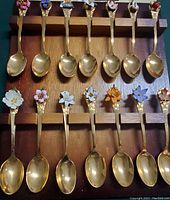 Photo of 14 gold plated collector spoons with floral enamel emblem handles displayed on a wooden rack showing color and detail of each spoon.