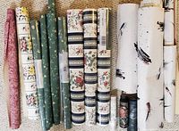 Multiple wallpaper rolls arranged upright, showing variety of floral and abstract patterns including green floral, white floral with stripes, and white with black brushstrokes