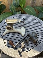 Two antique meat grinders placed on a wooden table outdoors with plants in background. One grinder has a wider base and tray, the other is more compact with a wooden handle.