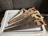 Four antique hand saws laid out on a plastic surface showing varying lengths and types of wooden handles, blades have rust and patina due to age.