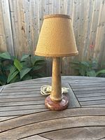 Table lamp with beige burlap shade and marble base displayed outdoors on a wooden table.