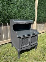 Black vintage style wooden nightstand shown outdoors on grass with a wooden fence and hedge background. The nightstand has a top shelf and a two-door cane inset cabinet below with ornate drawer handle and curved cabriole legs.
