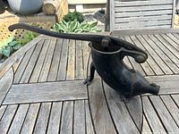 Side view of the vintage cast iron sausage stuffer showing the curved body, spout, and lever handle resting on a wooden table outdoors.
