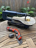 Mastercraft scroll saw and Black & Decker cordless reciprocating saw with charger on wooden table outdoors with plants in background