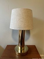 Front view of the table lamp showing the polished metal cylindrical base and beige fabric shade against a plain wall.