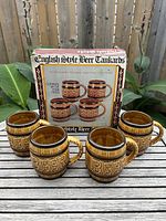 Set of five vintage ceramic beer mugs shown outside with original packaging behind them. Mugs have textured 'BEER' lettering and rustic glaze finish.