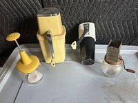 Photo showing a set of four vintage kitchen appliances: two ice crushers, one food chopper, and one meat chopper, on a black quilted background.