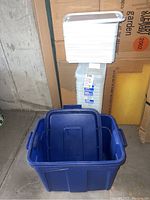 Blue plastic tote bin open and empty with stack of 29 plastic storage containers with white lids behind it.