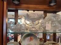 Shelf display of several cut glass serveware pieces including a large footed bowl, a rectangular dish, a small handled bowl, and a round platter in the background.