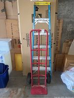 Front view of a red metal dolly/hand truck positioned in front of a 6-foot Werner ladder in a garage setting.