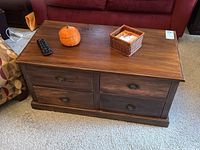 Wooden coffee table with four drawers, medium brown finish, decorative pumpkin and remote on top.