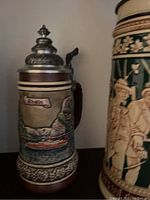 Photo showing two German beer steins, one with 'Rhein' relief and another green stein with figures raising glasses