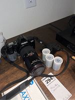 Minolta Maxxum 3000i camera with lens attached, camera strap, and three rolls of film on wooden surface, with some packaging visible.