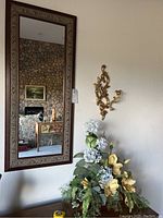 Tall rectangular mirror with decorative wooden frame, ornate gold-tone wall sconce mounted on adjacent wall, and potted flower arrangement with white and light yellow flowers.