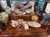 Photo of assorted handmade and collectible items arranged on a mirrored shelf including vases, lace items, ceramic dish, and a ceramic cottage music box.