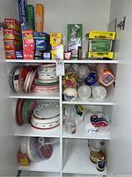 Photo showing white shelving unit with various boxes of Reynolds Wrap aluminum foil, Glad cling wrap, Ziploc bags, Hefty trash bags on top shelf, along with stacks of paper plates and foam cups on other shelves. Items are visibly heavily used and stored in a basement laundry room.